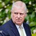 A close-up image of Andrew Mountbatten-Windsor. He is an older man with short white hair. He is wearing a dark blue suit jacket, a white shirt and a pale green and yellow patterned tie. He is looking away from the camera with a neutral expression on his face.