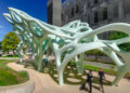 Outdoor public art installation featuring a large, pale green sculptural canopy with flowing, organic forms and cutout openings. The structure is supported by multiple curved legs and casts patterned shadows on the ground below. People walk beneath the sculpture along a landscaped pathway, with modern buildings and trees visible in the background under a clear blue sky.