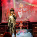 A performer dressed in a theatrical outfit with a top hat and makeup stands on stage, holding a microphone. Behind them is a large screen displaying dramatic imagery related to the performance, with the words 'Alice Cooper' written in a stylized, blood-like font. A drummer can be seen in the background.