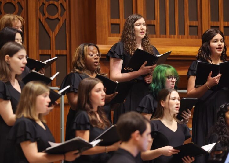 A choir of mostly women dressed in black hold music folders and sing in a campus chapel. Belle Voci
