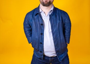 Bearded, light-skinned man with dwarfism stands smiling against a yellow portrait backdrop. He wears jeans, sneakers, a polo shirt, a jean jacket and a blue cap.
