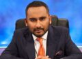 Amol Rajan wearing a blue suit and orange tie, sat on a chair and sat forward looking Ahead.