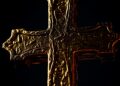 An extreme close-up photograph of a glowing, textured cross made of gold and bronze, using dramatic high-contrast studio lighting to create a sense of sacred, reverent glamour.