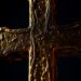 An extreme close-up photograph of a glowing, textured cross made of gold and bronze, using dramatic high-contrast studio lighting to create a sense of sacred, reverent glamour.