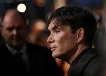 Cillian Murphy is pictured side-on, he is wearing a dark-coloured jacket and a dark-coloured shirt. Only his head and shoulders are seen. There are other people in the background, out of focus.