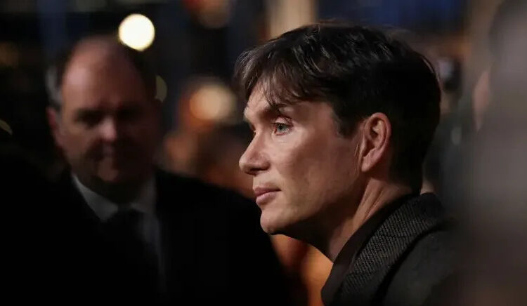 Cillian Murphy is pictured side-on, he is wearing a dark-coloured jacket and a dark-coloured shirt. Only his head and shoulders are seen. There are other people in the background, out of focus.