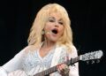 Dolly Parton wearing a sheer white blouse with balloon sleeves and white trousers holding a white and silver acoustic guitar on stage singing into a microphone attached to her head.