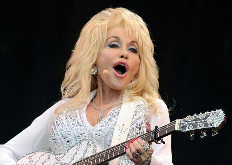Dolly Parton wearing a sheer white blouse with balloon sleeves and white trousers holding a white and silver acoustic guitar on stage singing into a microphone attached to her head.