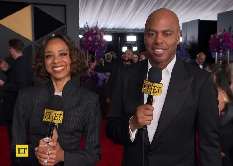 A woman and a man in black clothing holding microphones on a red carpet.