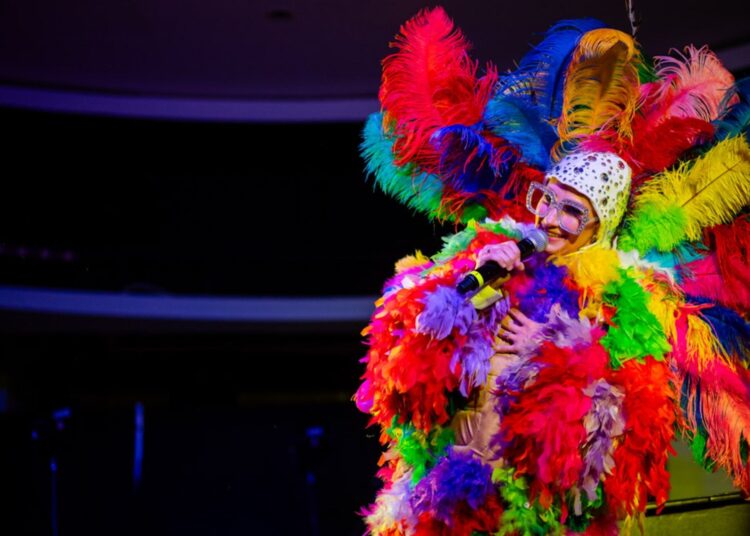 An Elton John tribute performer on stage covered in colorful feathers and big glasses