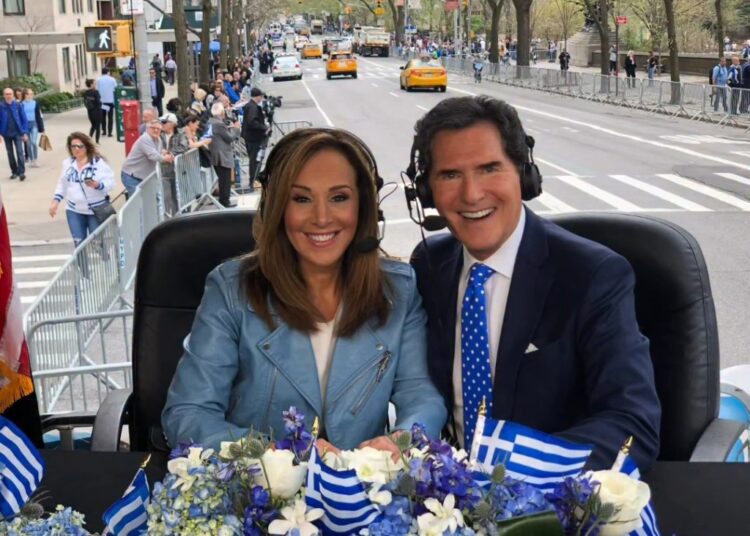 Two news anchors with headsets sit behind a table decorated with Greek flags and flowers, with a parade in the background.