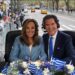 Two news anchors with headsets sit behind a table decorated with Greek flags and flowers, with a parade in the background.