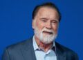 Arnold Schwarzenegger, an older man with black hair and a white beard standing smiling. He is wearing a blue shirt and dark jacket. The background is blue.