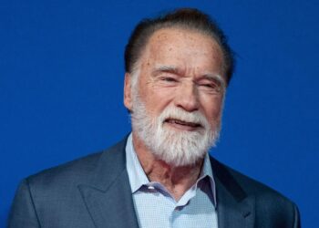 Arnold Schwarzenegger, an older man with black hair and a white beard standing smiling. He is wearing a blue shirt and dark jacket. The background is blue.