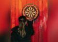 A woman with long dark blonde hair holds dart in front of a dartboard. The dartboard surround is orange and black and the dartboard is mounted on a wood panelling which is painted a rustic red. The woman is photographed from behind.