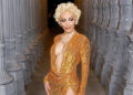 Doja Cat in a shimmering gold dress with short curly blonde hair poses between ornate columns.