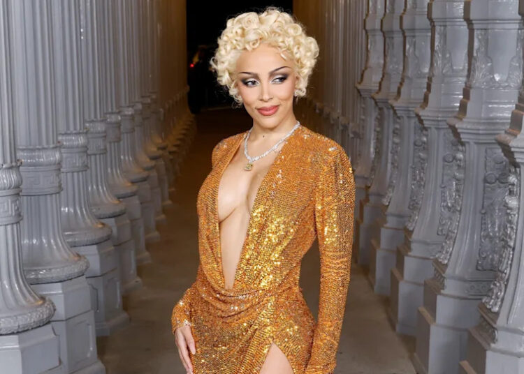 Doja Cat in a shimmering gold dress with short curly blonde hair poses between ornate columns.