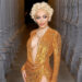 Doja Cat in a shimmering gold dress with short curly blonde hair poses between ornate columns.