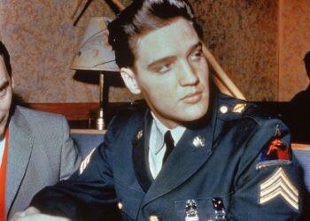 Elvis Presley with his hair in a neat quiff, wearing a military uniform. He appears to be sitting in a restaurant and is looking over his shoulder.