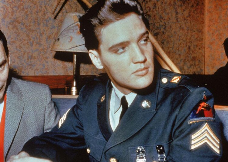 Elvis Presley with his hair in a neat quiff, wearing a military uniform. He appears to be sitting in a restaurant and is looking over his shoulder.