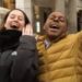 Two individuals standing outdoors in a public space. The individual on the left, dressed in a dark jacket, appears to be laughing and has their hands raised, while the person on the right, wearing a yellow jacket, has their arms raised and is also smiling. Behind them, there are architectural elements and a partially visible umbrella.