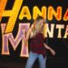 A woman with blond hair in a halter top and jeans looks downward as she stands in front of a "Hannah Montana" light display.