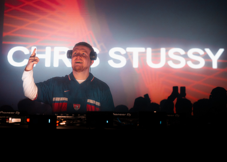 A DJ wearing headphones and a dark jersey points upward while smiling behind a Pioneer DJ booth, with “CHRIS STUSSY” displayed brightly in the background.