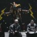 Two people sit on a stage while someone in a costume inspired by Indigenous cultural dress dances behind them.