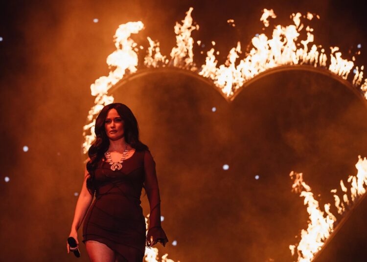 Kacey Musgraves stands on stage in a black dress in front of the shape of a heart on fire