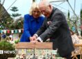 A picture of the King and Queen struggling to cut a cake at the Eden Project. The Queen is wearing a blue dress, the king has a grey suit.