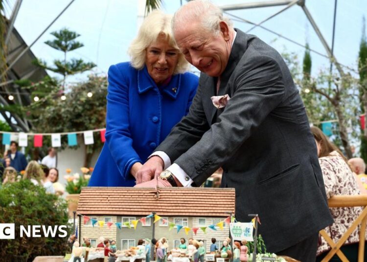 A picture of the King and Queen struggling to cut a cake at the Eden Project. The Queen is wearing a blue dress, the king has a grey suit.