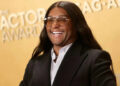 Person smiling on red carpet in sleek black suit and aviator glasses, in front of an awards backdrop