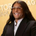 Person smiling on red carpet in sleek black suit and aviator glasses, in front of an awards backdrop