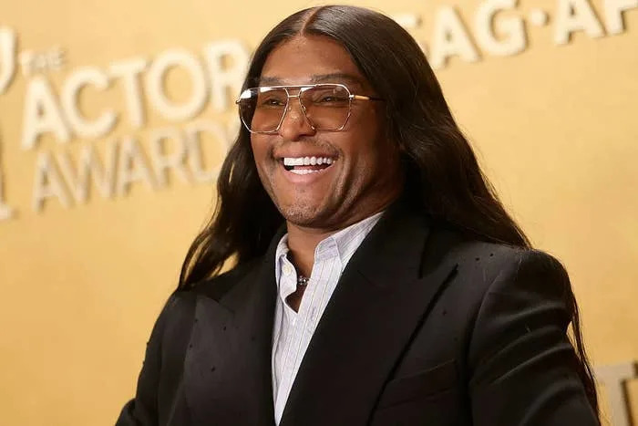 Person smiling on red carpet in sleek black suit and aviator glasses, in front of an awards backdrop