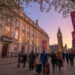 “a cinematic wide-angle view of smith square hall in london during early spring evening, warm golden lights glowing from inside the historic concert hall, elegantly dressed international audience arriving, big ben and the london skyline softly visible in the background, musicians carrying violins and cellos at the entrance, banners reading ‘london festival of chamber music 2026’, cherry blossoms beginning to bloom, атмосферic twilight sky, ultra-detailed, realistic, vibrant colors, travel magazine style photography, 8k resolution