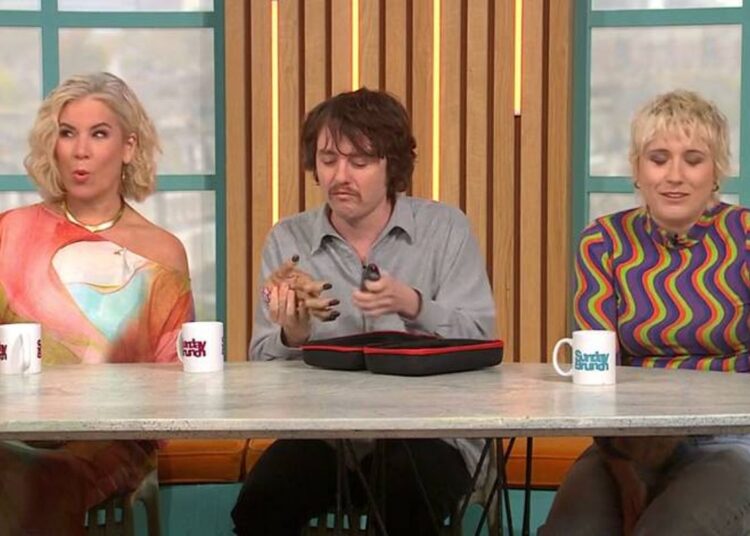 Three individuals are seated at a table, possibly engaged in a conversation or discussion. Each person is holding a cup in their hand. The table is adorned with a tray and additional cups. The backdrop features a striped wall and a window.