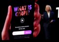 A woman in a dark pantsuit stands presenting to an audience off camera. behind her, a big screen displays a phone in hand, and hte display on the phone reads "what is quibi?"