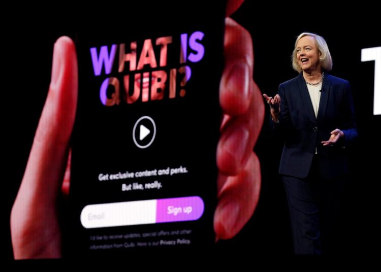 A woman in a dark pantsuit stands presenting to an audience off camera. behind her, a big screen displays a phone in hand, and hte display on the phone reads "what is quibi?"