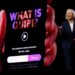 A woman in a dark pantsuit stands presenting to an audience off camera. behind her, a big screen displays a phone in hand, and hte display on the phone reads "what is quibi?"