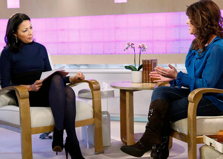 Two women, including Amy Duggar, sitting in chairs having a conversation in a studio setting with a pink-lit background.