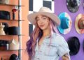 A woman with long hair which is dyed pink at the front, purple towards the back, looks towards the camera while wearing a cream, wide-brimmed hat. She wears a light blue short-sleeved denim top. Behind her are a variety of hats hanging on the wall or on shelves.
