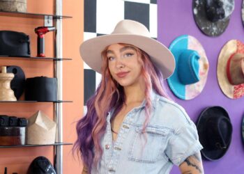 A woman with long hair which is dyed pink at the front, purple towards the back, looks towards the camera while wearing a cream, wide-brimmed hat. She wears a light blue short-sleeved denim top. Behind her are a variety of hats hanging on the wall or on shelves.
