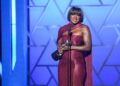 A woman in a purple dress holding a trophy in her hands standing at a microphone onstage.