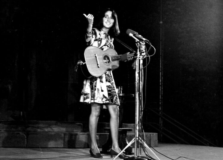a woman in a knee-length dress stands in front of a microphone holding a guitar on stage, speaking to the crowd and pointing out