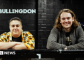 Hannah Bruce and James Taylor are leaning on a black metal bar. She is on the left wearing a flowery short and has short hair. He is on the right, has long hair and is wearing a grey jumper. Behind them is a black wall with 'The Bullingdon' in white writing.