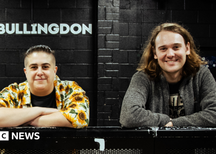 Hannah Bruce and James Taylor are leaning on a black metal bar. She is on the left wearing a flowery short and has short hair. He is on the right, has long hair and is wearing a grey jumper. Behind them is a black wall with 'The Bullingdon' in white writing.