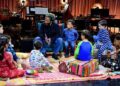 Musician Ziggy Marley sits on a chair holding a book while a group of young children in pajamas gather around him on colorful rugs and cushions. The children sit in a semicircle, listening attentively. Behind them is a stage setup with musical instruments, including drums, keyboards, and stands, softly lit by warm studio lights.