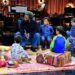 Musician Ziggy Marley sits on a chair holding a book while a group of young children in pajamas gather around him on colorful rugs and cushions. The children sit in a semicircle, listening attentively. Behind them is a stage setup with musical instruments, including drums, keyboards, and stands, softly lit by warm studio lights.