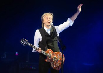 Paul McCartney performing on stage with a red guitar and one arm raised.