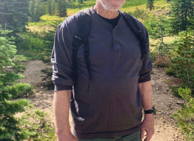 Chuck Norris wearing a black cap, gray shirt, and green shorts, standing on a hiking trail.